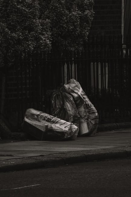 Two large black plastic bags filled with waste materials are positioned on the pavement near the edge of a road, leaning slightly against a black metal fence with vertical bars. One bag appears to contain crumpled paper or cardboard, while the other has a less defined mix of waste, possibly plastic or fabric items. The surface of the bags shows creases and reflections, indicating they are made of glossy, durable plastic. In the background, dense foliage of leafy green trees is visible, partially obscuring a brick building behind the fence. The scene is outdoors, with natural lighting casting soft shadows on the ground and bags, suggesting it is daytime. The setting appears to relate to private waste collection or rubbish removal services, and is consistent with the context of alternative rubbish clearance methods outside of municipal collection, as offered by companies such as Rubbish Clearance Wandsworth.