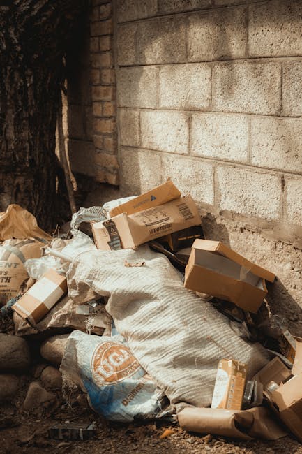 Two large black plastic bags filled with waste materials are positioned on the pavement near the edge of a road, leaning slightly against a black metal fence with vertical bars. One bag appears to contain crumpled paper or cardboard, while the other has a less defined mix of waste, possibly plastic or fabric items. The surface of the bags shows creases and reflections, indicating they are made of glossy, durable plastic. In the background, dense foliage of leafy green trees is visible, partially obscuring a brick building behind the fence. The scene is outdoors, with natural lighting casting soft shadows on the ground and bags, suggesting it is daytime. The setting appears to relate to private waste collection or rubbish removal services, and is consistent with the context of alternative rubbish clearance methods outside of municipal collection, as offered by companies such as Rubbish Clearance Wandsworth.