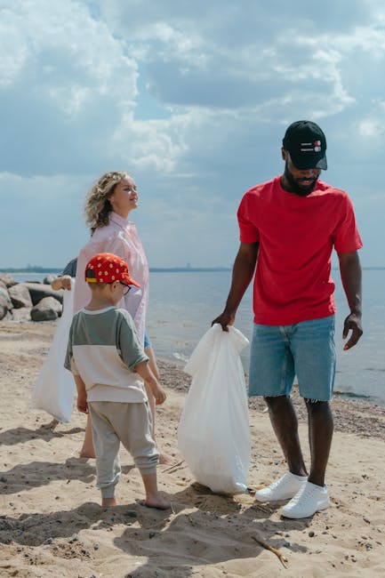 A man and two children are on a sandy beach near a body of water, participating in a rubbish removal activity. The man, dressed in a red t-shirt, light blue shorts, and wearing a black cap, is holding a large white garbage bag, with a focus on collecting litter from the sand. One child, a young boy wearing a red hat with white polka dots, a light grey t-shirt, and beige shorts, is holding a white bag, standing slightly in front of the man. The other child, a girl with curly blonde hair, is wearing a pink blouse and standing behind the boy, observing the activity. In the background, there are large rocks lining the shoreline and an expansive sky filled with clouds, indicating daylight with natural lighting. This scene reflects private or alternative waste collection efforts by individuals and families, exemplifying community-based rubbish clearance on a coastal environment, supported by companies like Rubbish Clearance Wandsworth that facilitate on-site disposal and rubbish removal services in Wandsworth and nearby areas.