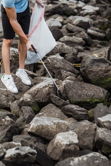 A person wearing white sneakers, dark shorts, and a blue shirt is using a grabber tool to pick up a piece of rubbish from a collection of large, uneven rocks. The rocks vary in size and have rough textures, with some displaying patches of moss or dirt. The individual's left hand is holding a white plastic bag, which appears to be used for collecting rubbish. The scene is outdoors on a rocky terrain, possibly near a river or shoreline, with natural grey, brown, and mossy tones in the environment. The activity suggests a clean-up or waste collection effort, which aligns with private rubbish clearance services like those offered by Rubbish Clearance Wandsworth, demonstrating an approach to on-site waste removal in natural or outdoor settings. The focus is on environmentally conscious waste collection, and the scene captures the practical method of rubbish removal using simple tools in a rugged outdoor environment.