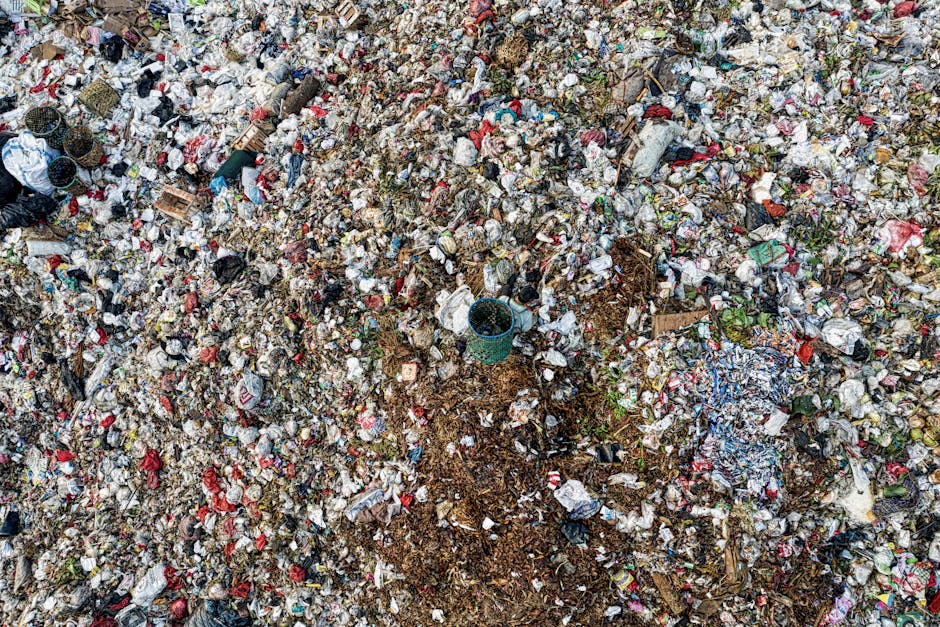 A large, uncontained pile of mixed waste and rubbish spans across an outdoor area, consisting of various materials such as plastic bottles, crushed paper, broken cardboard, and discarded packaging in multiple colors, predominantly white, red, and blue. The texture of the waste appears crumpled, torn, and scattered, with some items partially buried under others. Amidst the debris, there are a few small, empty plastic containers and tangled plastic bags, some of which are visibly weathered and dirty. The waste occupies the majority of the scene, with a section of bare, compacted soil or earth visible in the middle, suggesting recent disturbance or partial clearing. The surrounding environment indicates an open, possibly neglected space, with no clearly defined boundary or containment visible in the image. The diffuse natural lighting highlights the chaos of the refuse, aligning with disposal challenges faced through private waste handling and alternative rubbish removal services such as those offered by Rubbish Clearance Wandsworth, which specializes in managing bulky waste and large-scale rubbish clearance for properties, construction sites, or land clearing purposes.