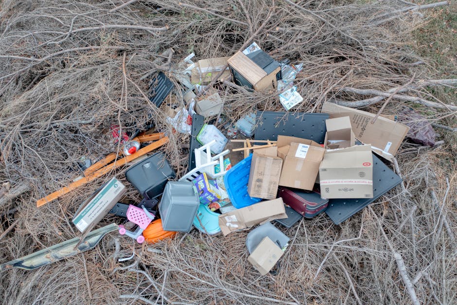 A large, uncontained pile of mixed waste and rubbish spans across an outdoor area, consisting of various materials such as plastic bottles, crushed paper, broken cardboard, and discarded packaging in multiple colors, predominantly white, red, and blue. The texture of the waste appears crumpled, torn, and scattered, with some items partially buried under others. Amidst the debris, there are a few small, empty plastic containers and tangled plastic bags, some of which are visibly weathered and dirty. The waste occupies the majority of the scene, with a section of bare, compacted soil or earth visible in the middle, suggesting recent disturbance or partial clearing. The surrounding environment indicates an open, possibly neglected space, with no clearly defined boundary or containment visible in the image. The diffuse natural lighting highlights the chaos of the refuse, aligning with disposal challenges faced through private waste handling and alternative rubbish removal services such as those offered by Rubbish Clearance Wandsworth, which specializes in managing bulky waste and large-scale rubbish clearance for properties, construction sites, or land clearing purposes.