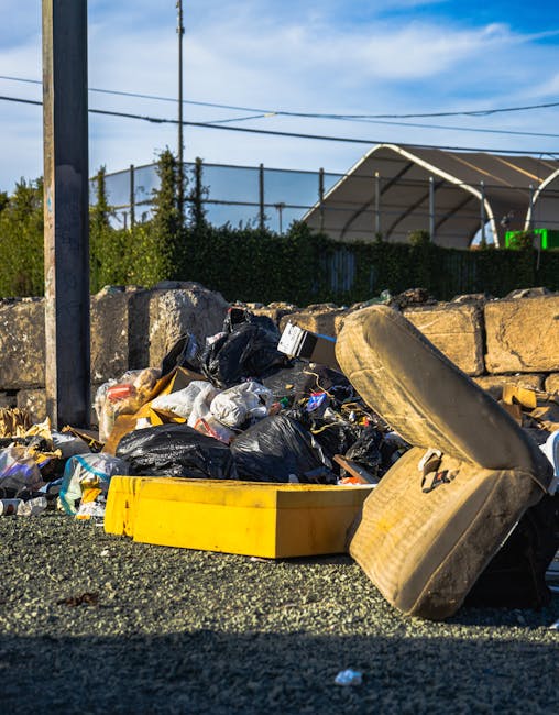 A close-up view of a pile of dumped rubbish on a gravel surface in an outdoor setting. The debris includes multiple black plastic garbage bags, partially torn, revealing assorted household waste inside. Among the rubbish, there is a yellow plastic container, possibly intended for storage or disposal, and an old, worn car tire leaning against the pile, with visible dirt and weathering. In the background, a low stone wall made of large, uneven stones partially encloses the area, and behind it, a green hedge and a sports netted fence are visible. The scene is illuminated by natural daylight, with a partly cloudy blue sky overhead, and utility poles with wires stretching across the scene. The image reflects a situation where independent collection or illegal dumping of waste has occurred in an outdoor environment, highlighting the importance of proper rubbish removal services such as those offered by Rubbish Clearance Wandsworth.